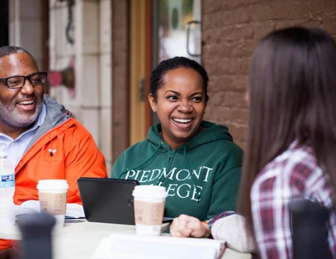 Three people sit at an outdoor table, smiling and talking with coffee cups in front of them. One woman wears a green “Piedmont College” hoodie, another has long brown hair, and a man wears glasses and an orange jacket.