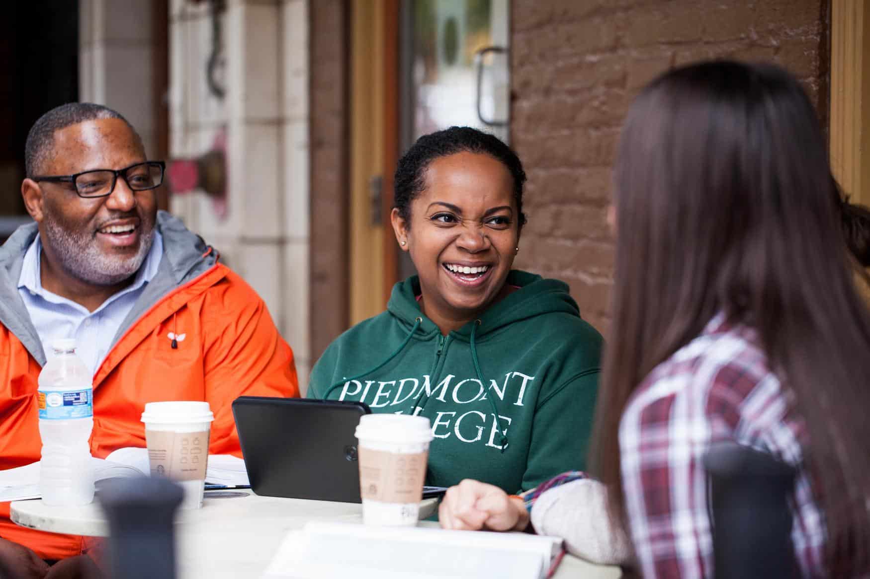 Three people sit at an outdoor table, smiling and talking with coffee cups in front of them. One woman wears a green “Piedmont College” hoodie, another has long brown hair, and a man wears glasses and an orange jacket.