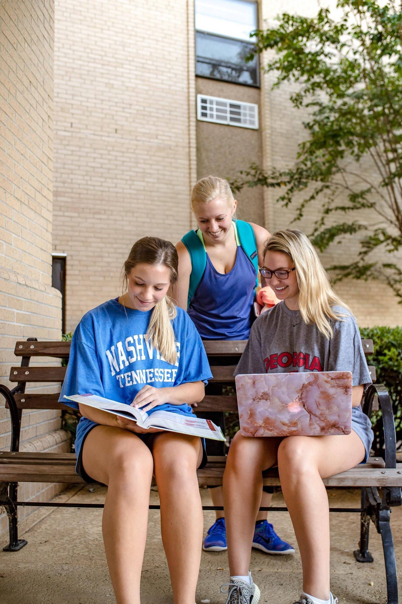 Three young women study outside on a bench. One reads a book, another uses a laptop, and the third stands behind them with a backpack and an apple, smiling. They are near a brick building and some greenery.