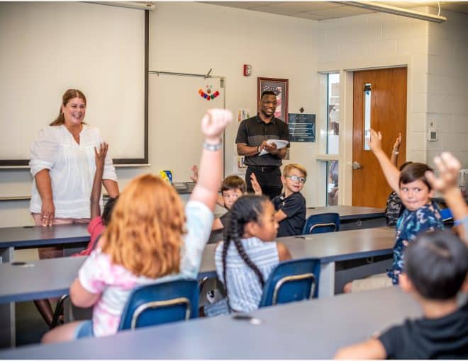 A classroom with students sitting at desks, raising their hands. A woman stands at the front smiling, while a man stands near the door holding a notebook, also smiling. A whiteboard and projector are visible behind them.