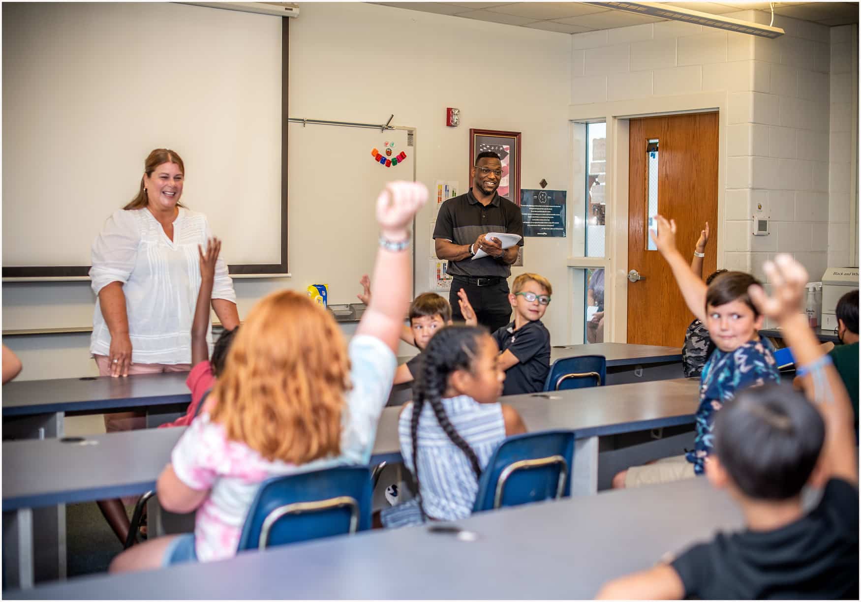 A classroom with students sitting at desks, raising their hands. A woman stands at the front smiling, while a man stands near the door holding a notebook, also smiling. A whiteboard and projector are visible behind them.