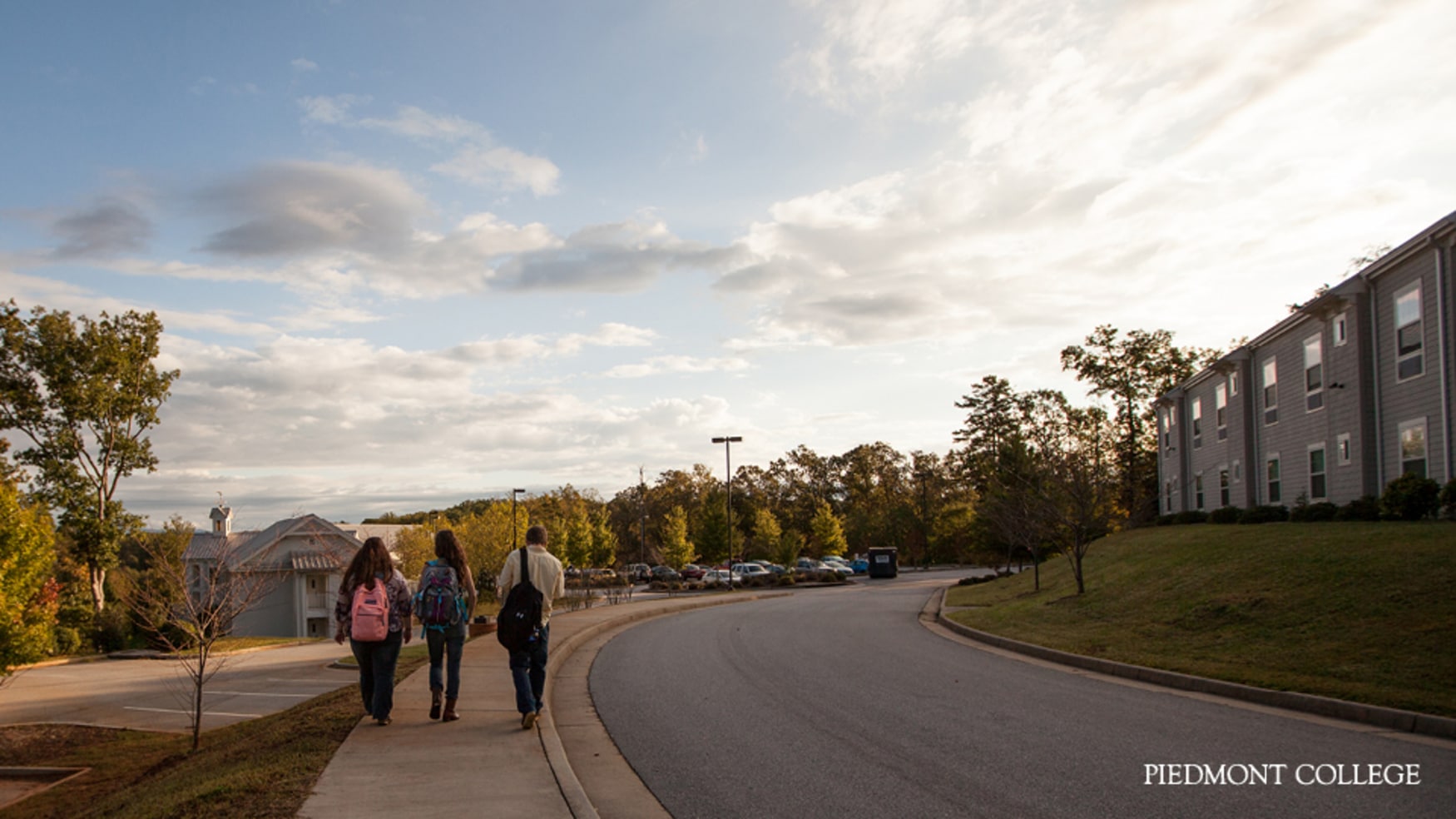 Three students walk along a curved sidewalk on a college campus, with trees, parked cars, and a building under a partly cloudy sky. The words Piedmont College appear in the bottom right corner.