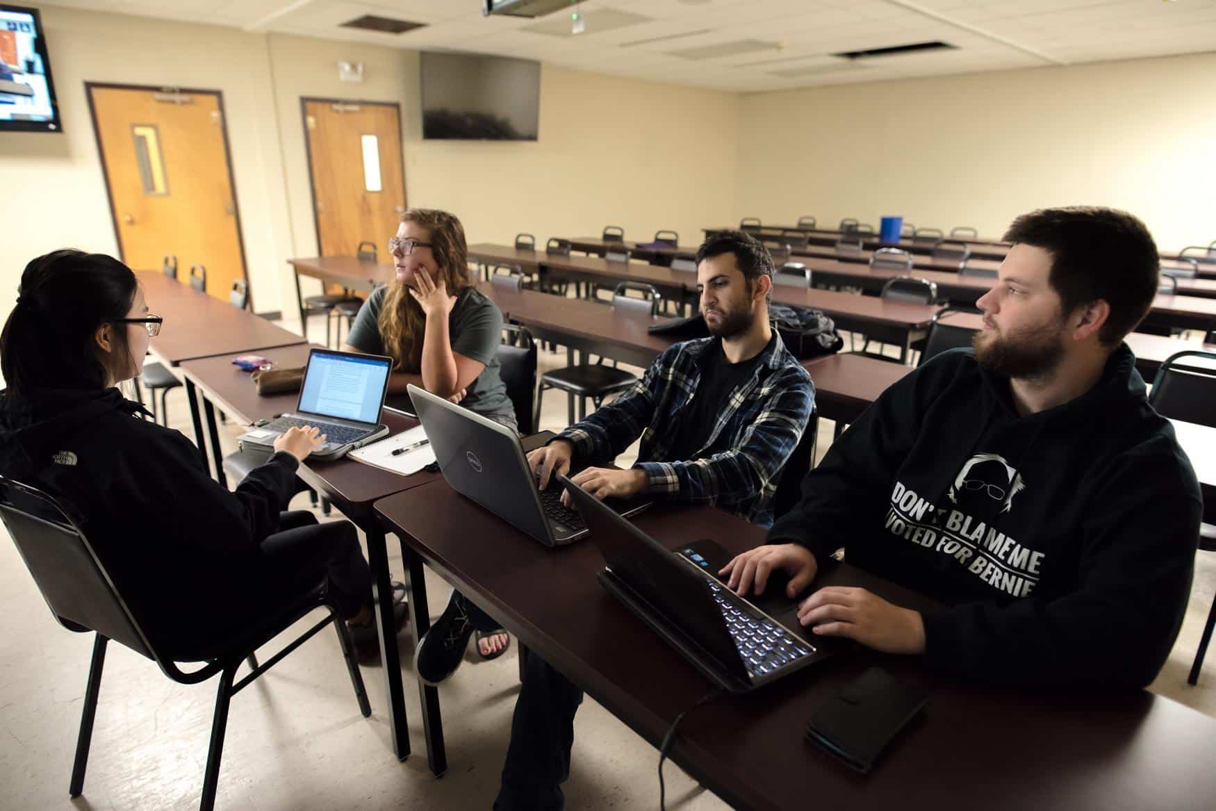 Four students with laptops sit in a classroom at narrow tables