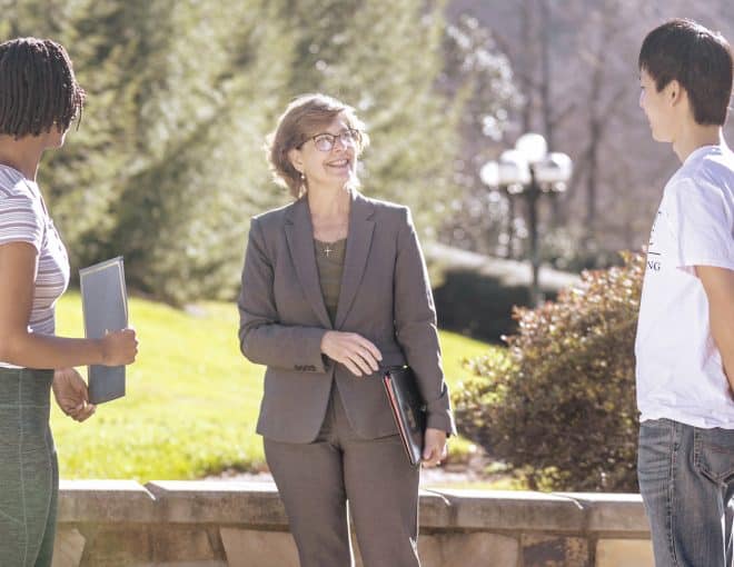 A woman in a suit stands outdoors, smiling and talking with two students, one holding a notebook. The group is near greenery on a sunny day.