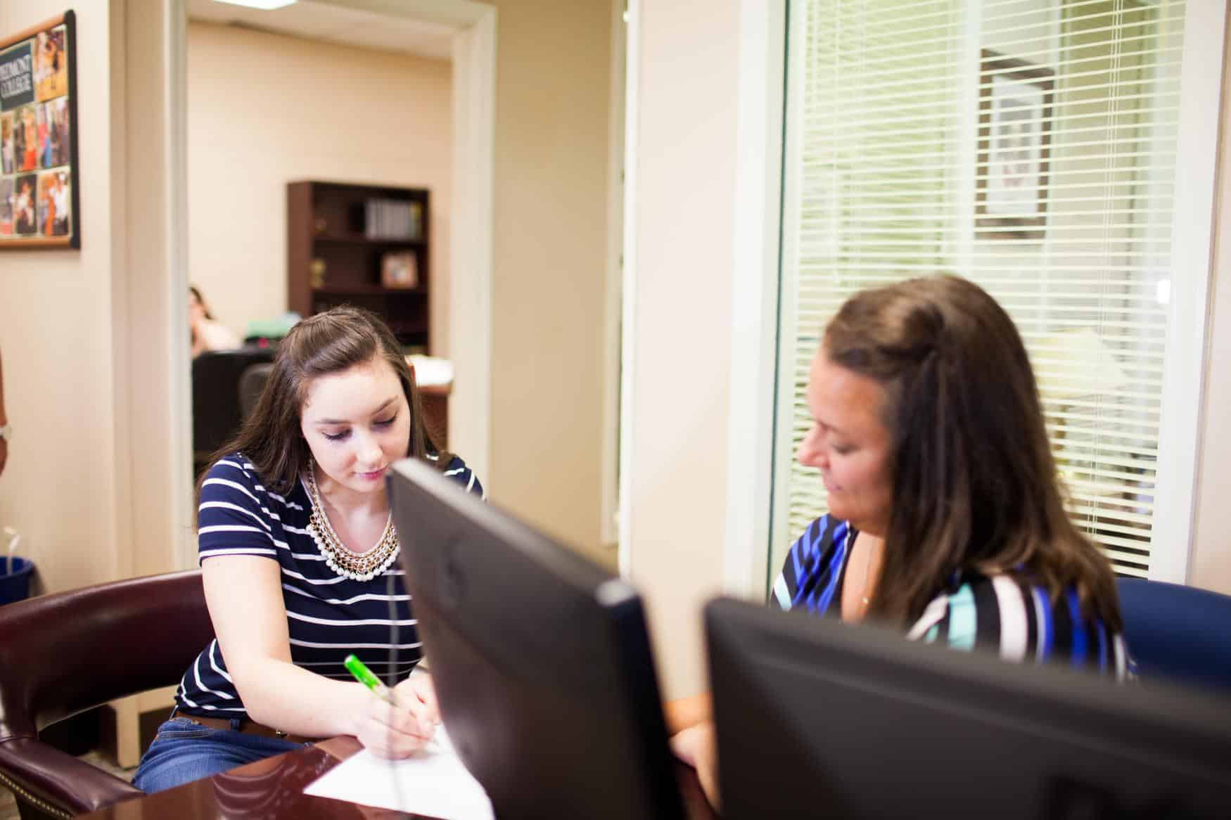 Two women sitting at a desk behind computer monitors, going over a document.