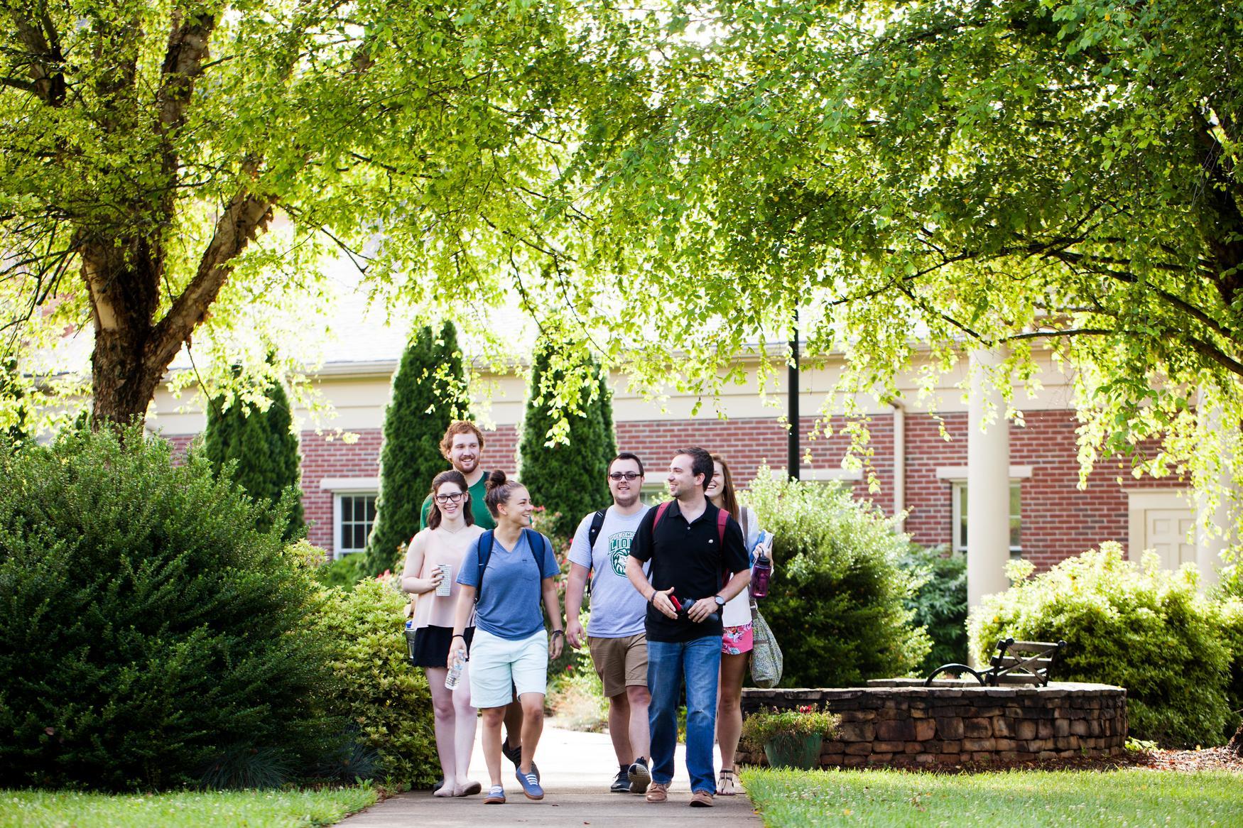 Group of students walking down a pathway. Green trees frame them, and a brick building is the background.