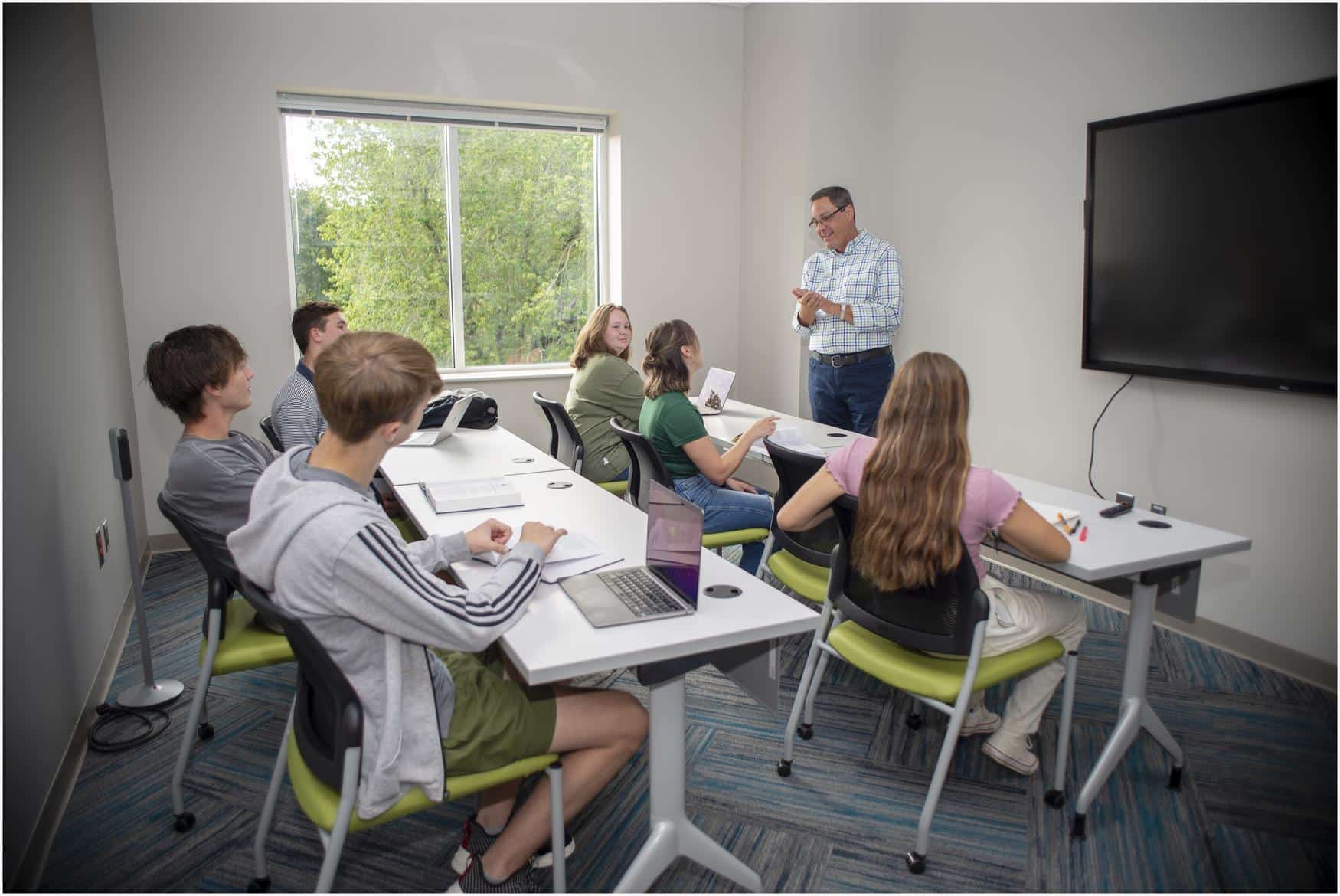 Professor teaching a class of six students. The classroom is bright, with trees visible through the window.