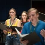 A group of students holding choir music look off camera while singing
