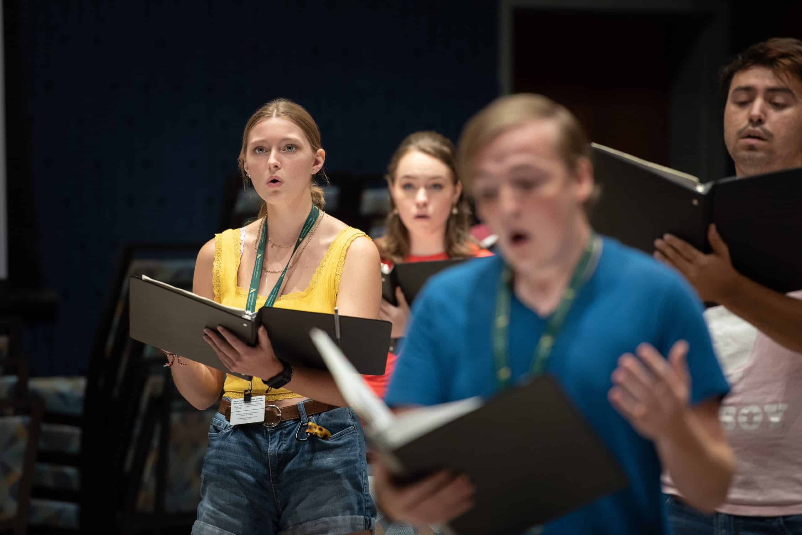 A group of students holding choir music look off camera while singing