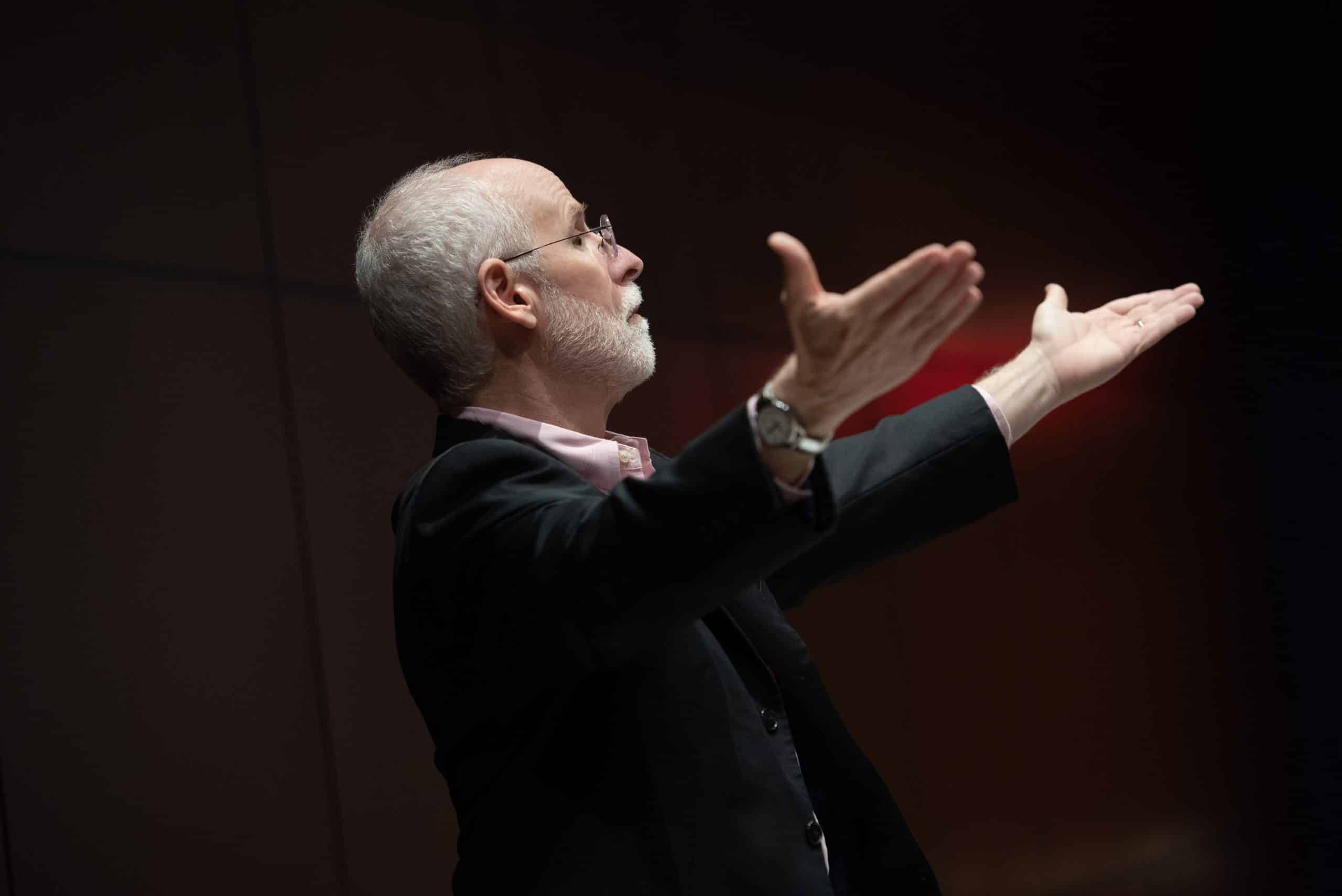 A conductor wearing a black suit stands in. profile, with his hands raised.