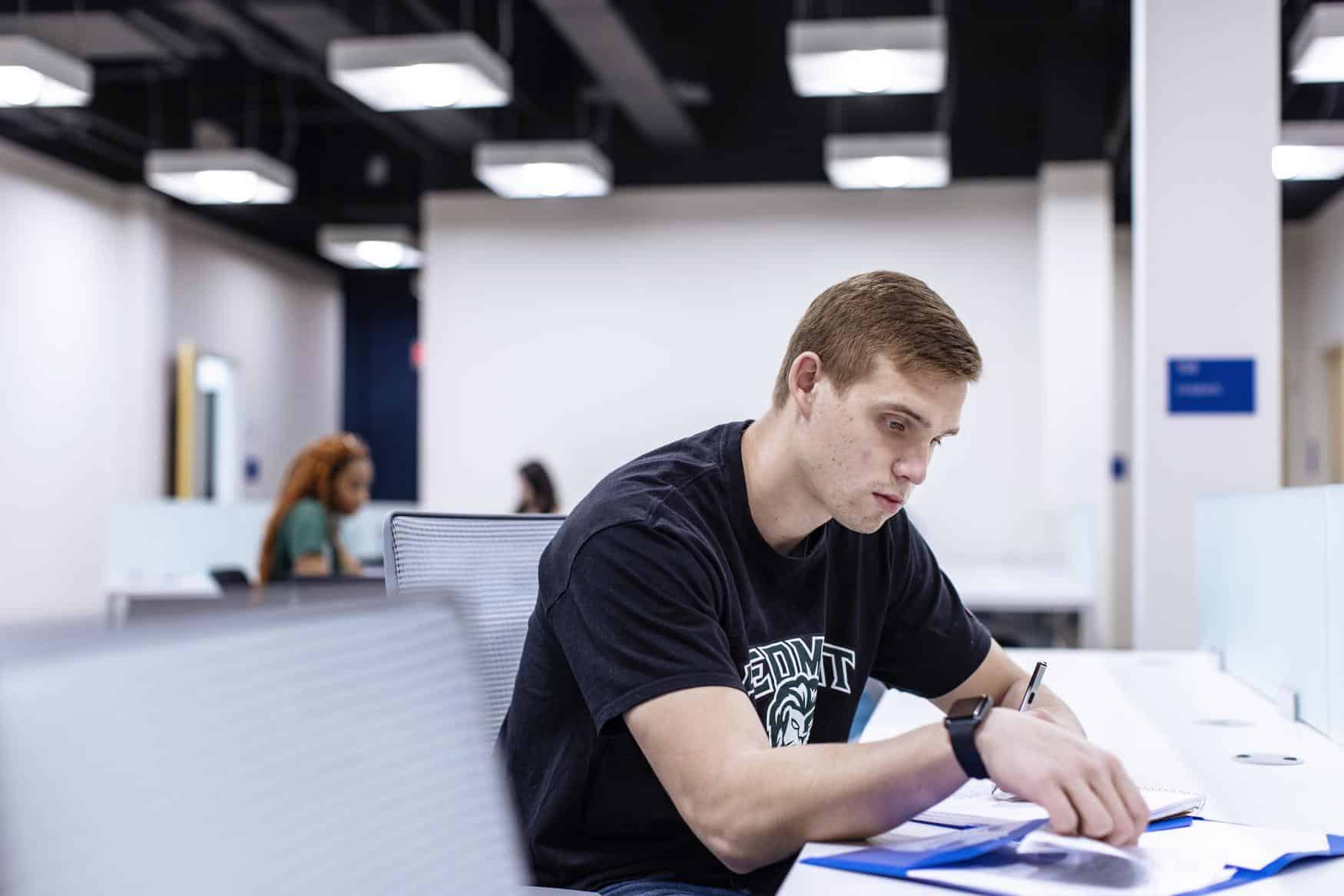 Young man wearing a Piedmont shirt leaning over a desk and studying papers. Other students are visible but blurred in the background.