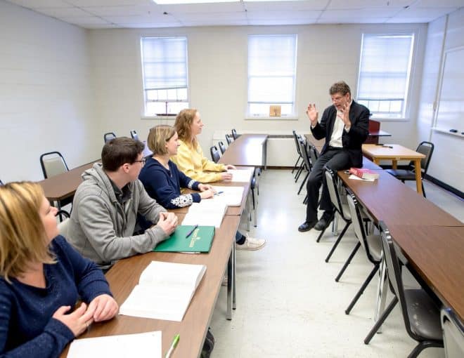 A professor leans against a table in class, speaking animatedly with his hands raised. A row of students seated at desks intently listen.