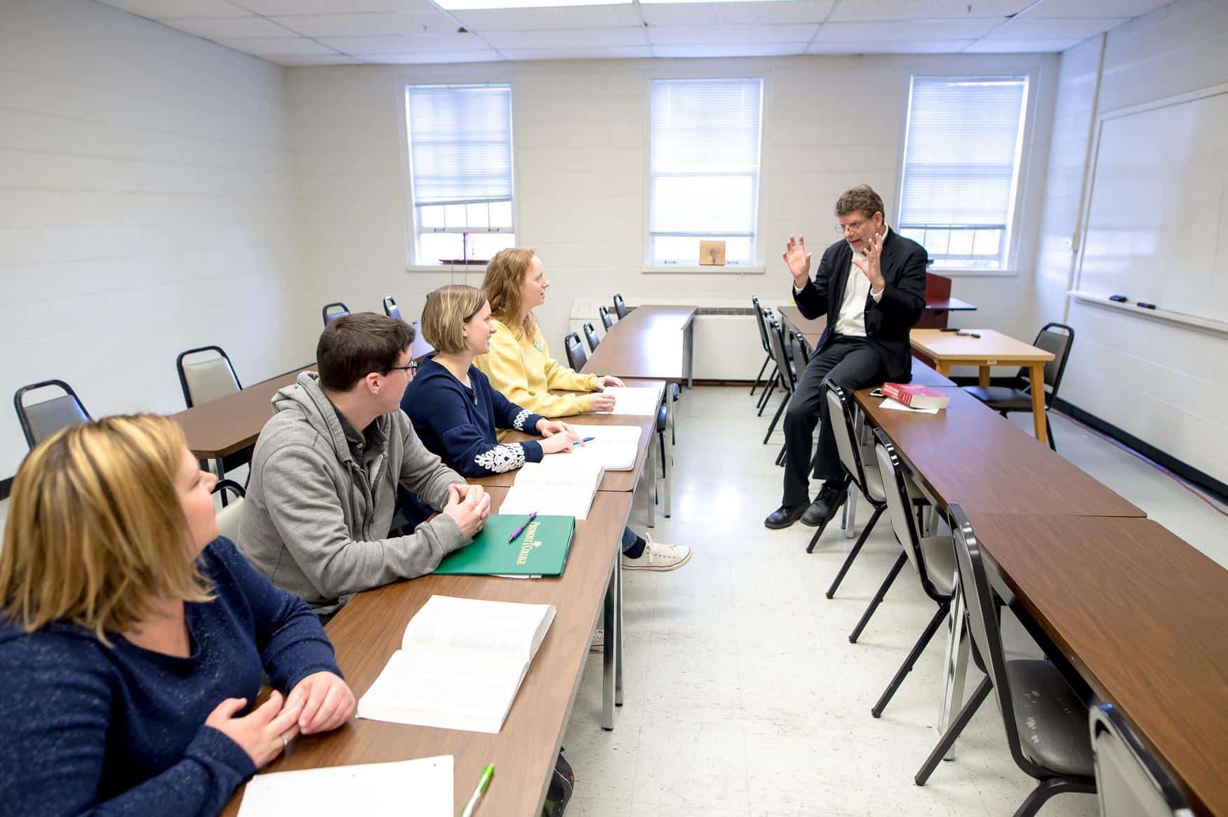 A professor leans against a table in class, speaking animatedly with his hands raised. A row of students seated at desks intently listen.