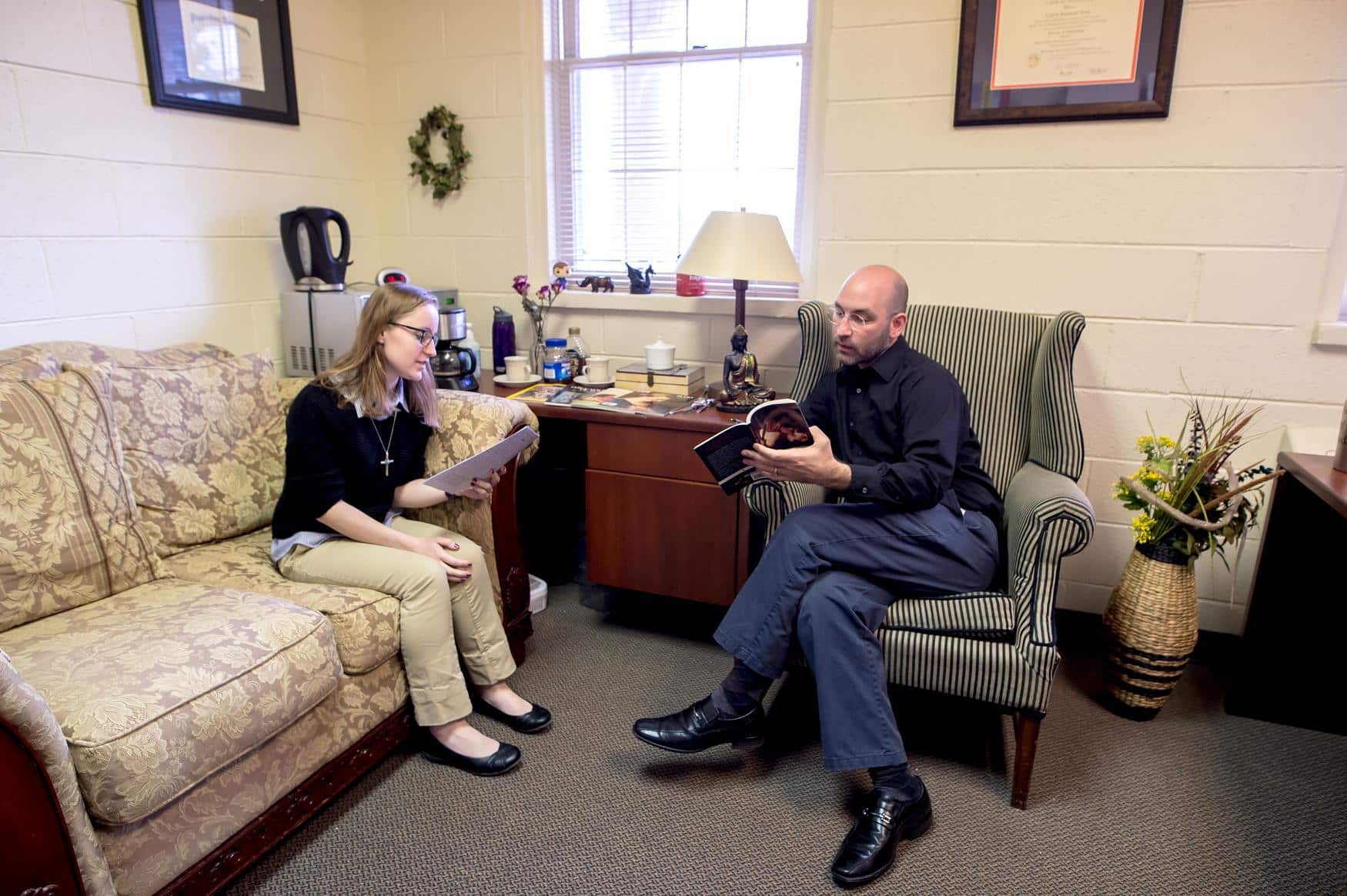 Two adults sitting on a sofa and chair, holding books and paper. They look to be mid-conversation