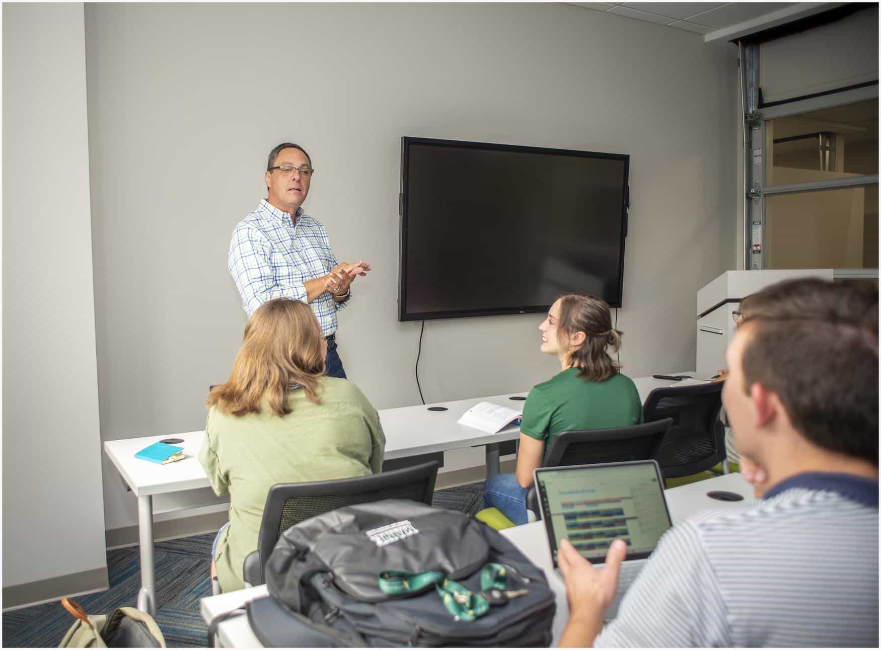 A professor stand in front of a class mid-lecture. Students face towards him and a television is visible on the wall in the background.