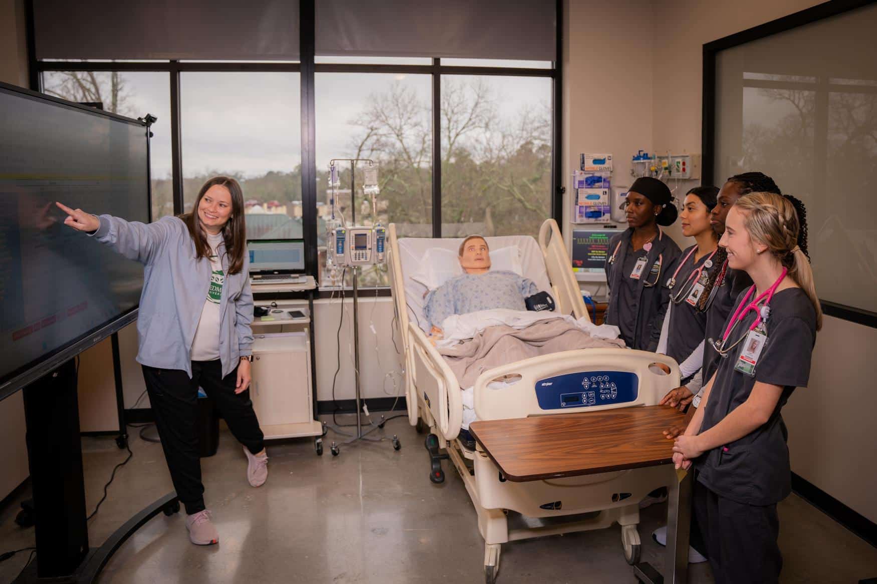 A group of nursing students stand behind a medical dummy in a hospital bed, watching an instructor across from them point to an interactive screen.