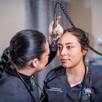 A student holds medical equipment up to another student, practicing an examination.