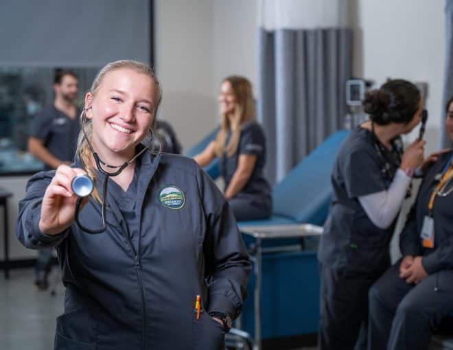 A nursing student smiles at the camera and points a stethoscope towards it. Other students are visible in the background.