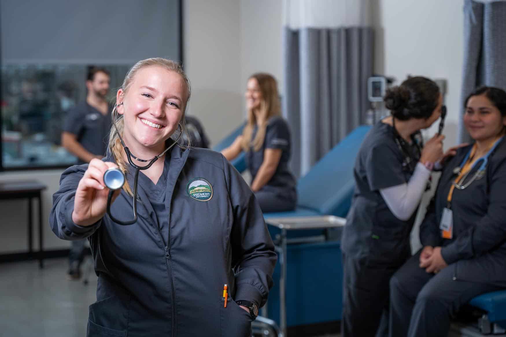 A nursing student smiles at the camera and points a stethoscope towards it. Other students are visible in the background.