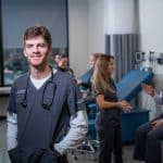 A young man in nursing scrubs smiles for the camera. Behind him, a group of students wearing scrubs can be seen in conversation.