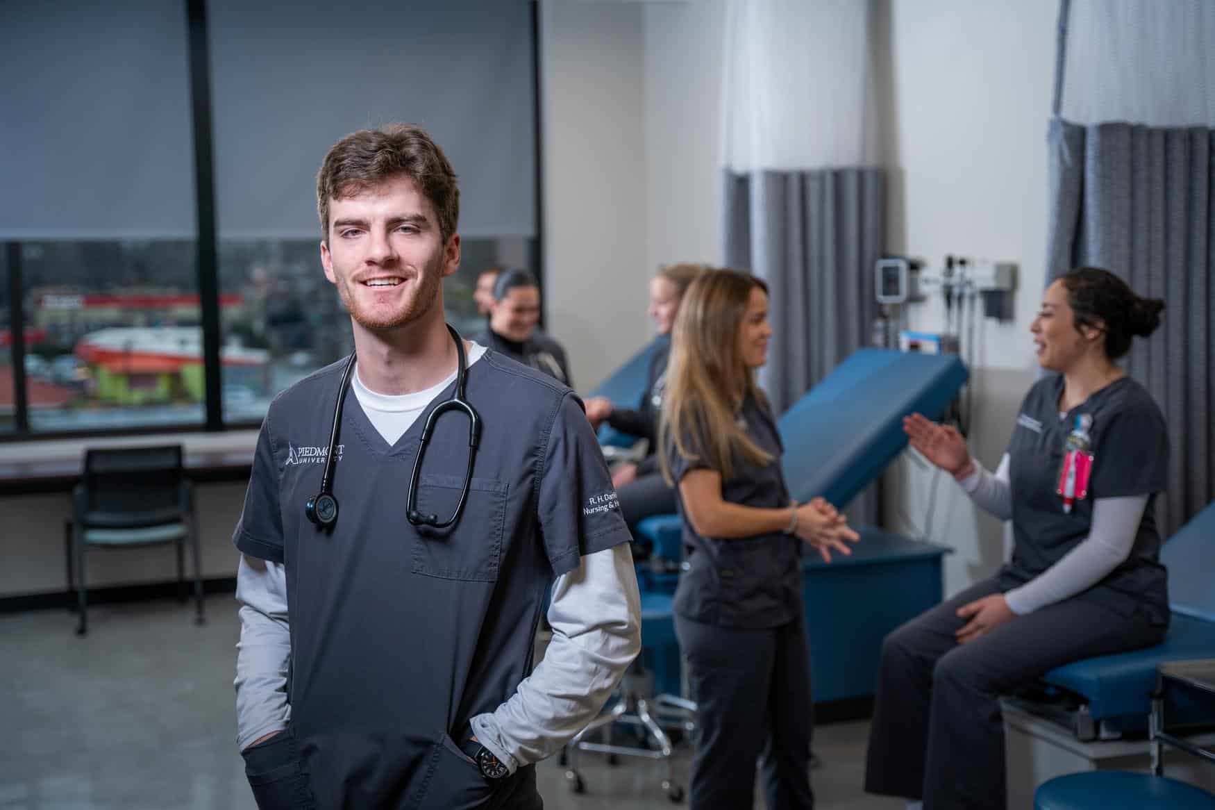 A young man in nursing scrubs smiles for the camera. Behind him, a group of students wearing scrubs can be seen in conversation.