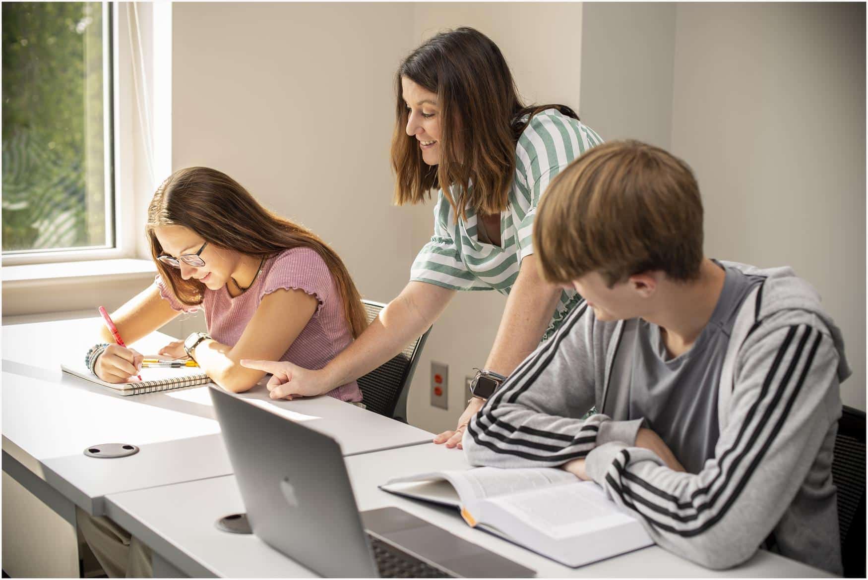 Three adults in a row at a table, in a classroom setting. One adult is taking notes, one is pointing to the notes, and the other is sitting with an open laptop and book.