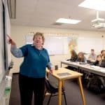 Professor pointing to a white board to the left of the image, with students blurred and visible in the right background.
