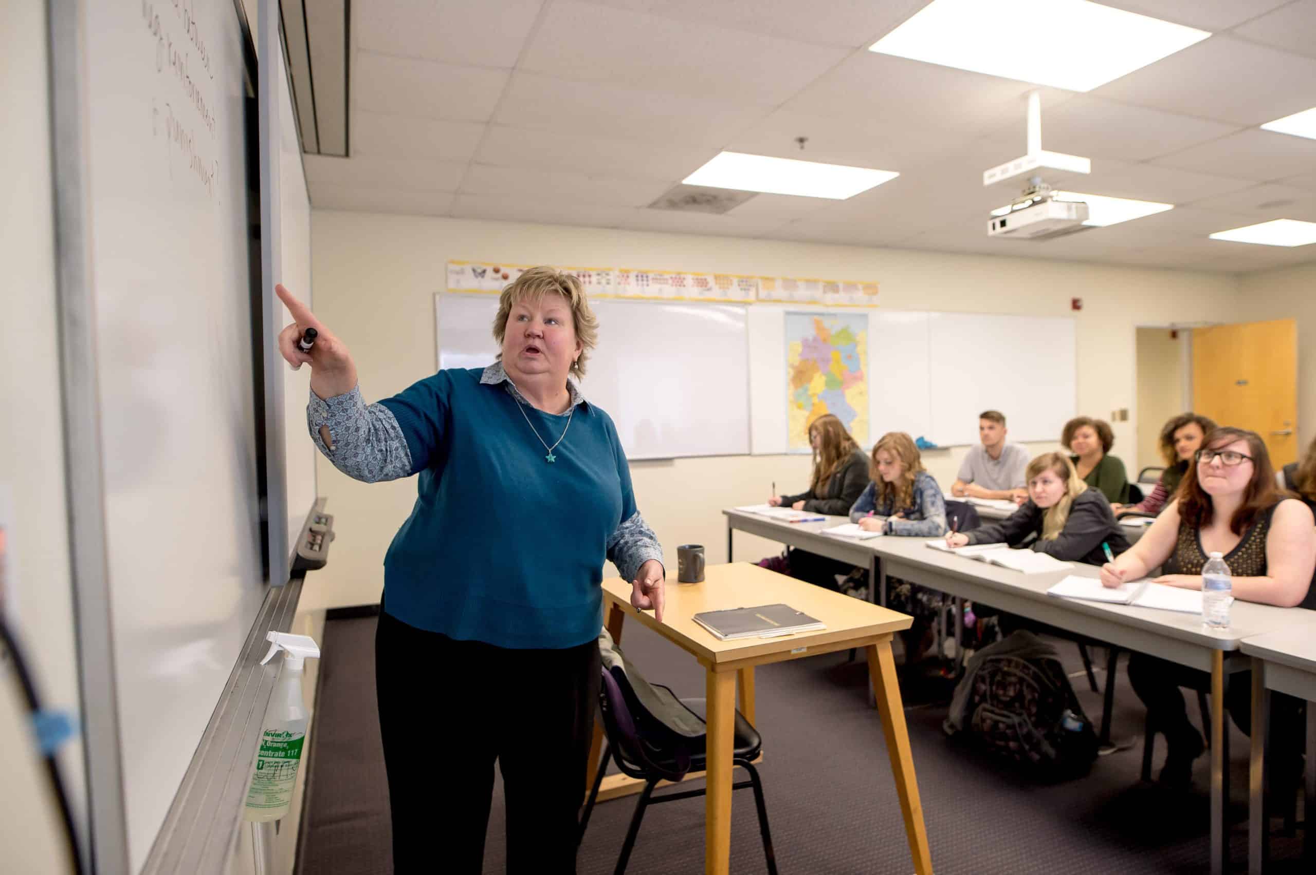 Professor pointing to a white board on the left, while her students watch.