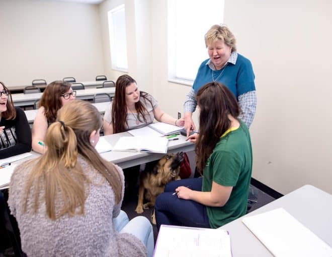A professor points and smiles to a service dog hidden under the classroom tables, while a group of students looks on
