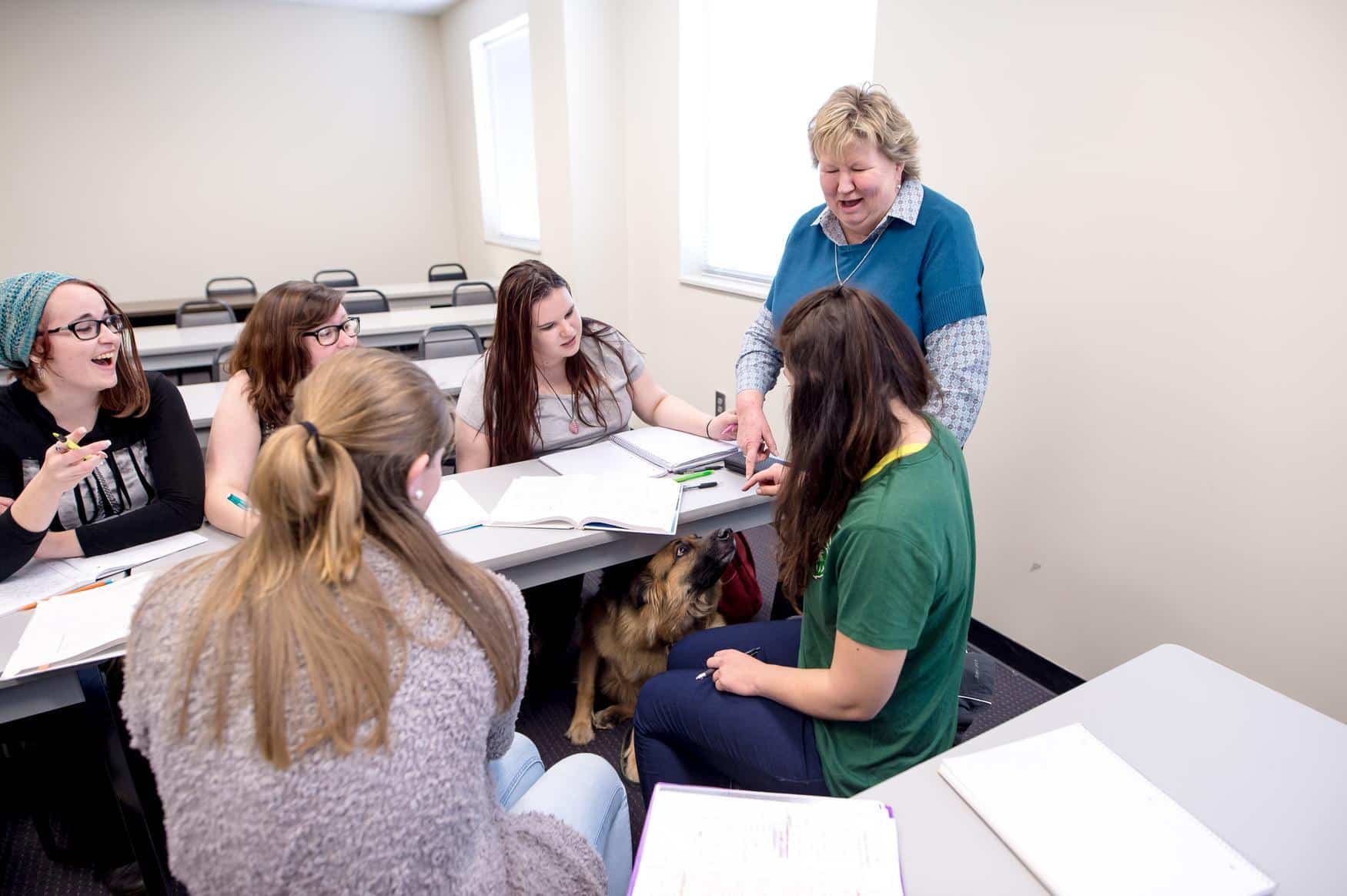 A professor points and smiles to a service dog hidden under the classroom tables, while a group of students looks on
