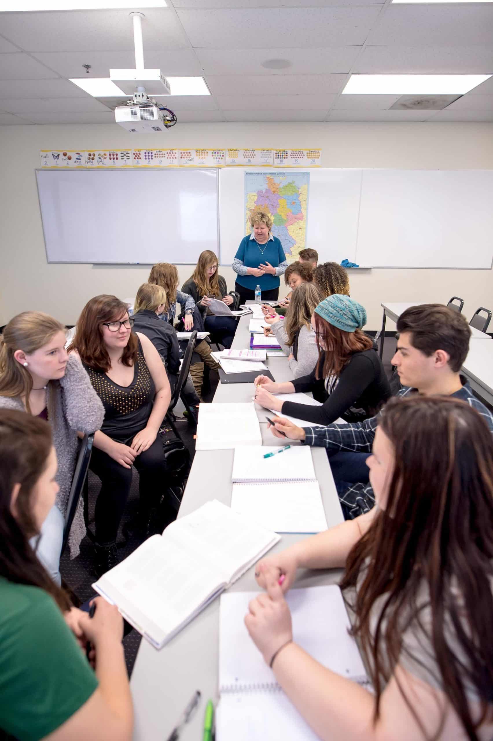 Vertical image of a group of students at a narrow table sitting across from one another. At the end of the table a professor stands, in conversation with a student.