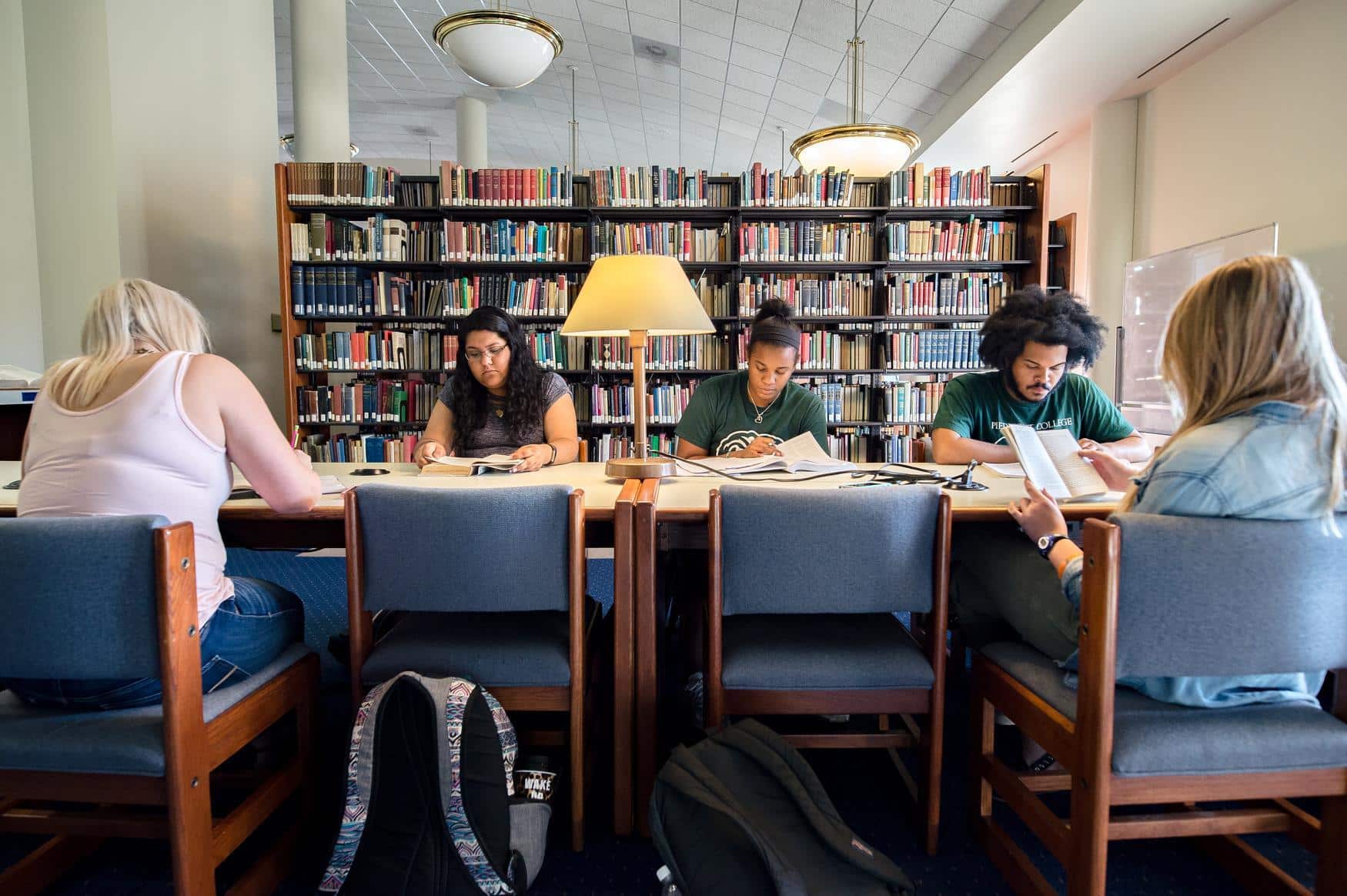 Group of students at a library table with a lamp on it. The students are reading books. Shelves of books are visible in the background.