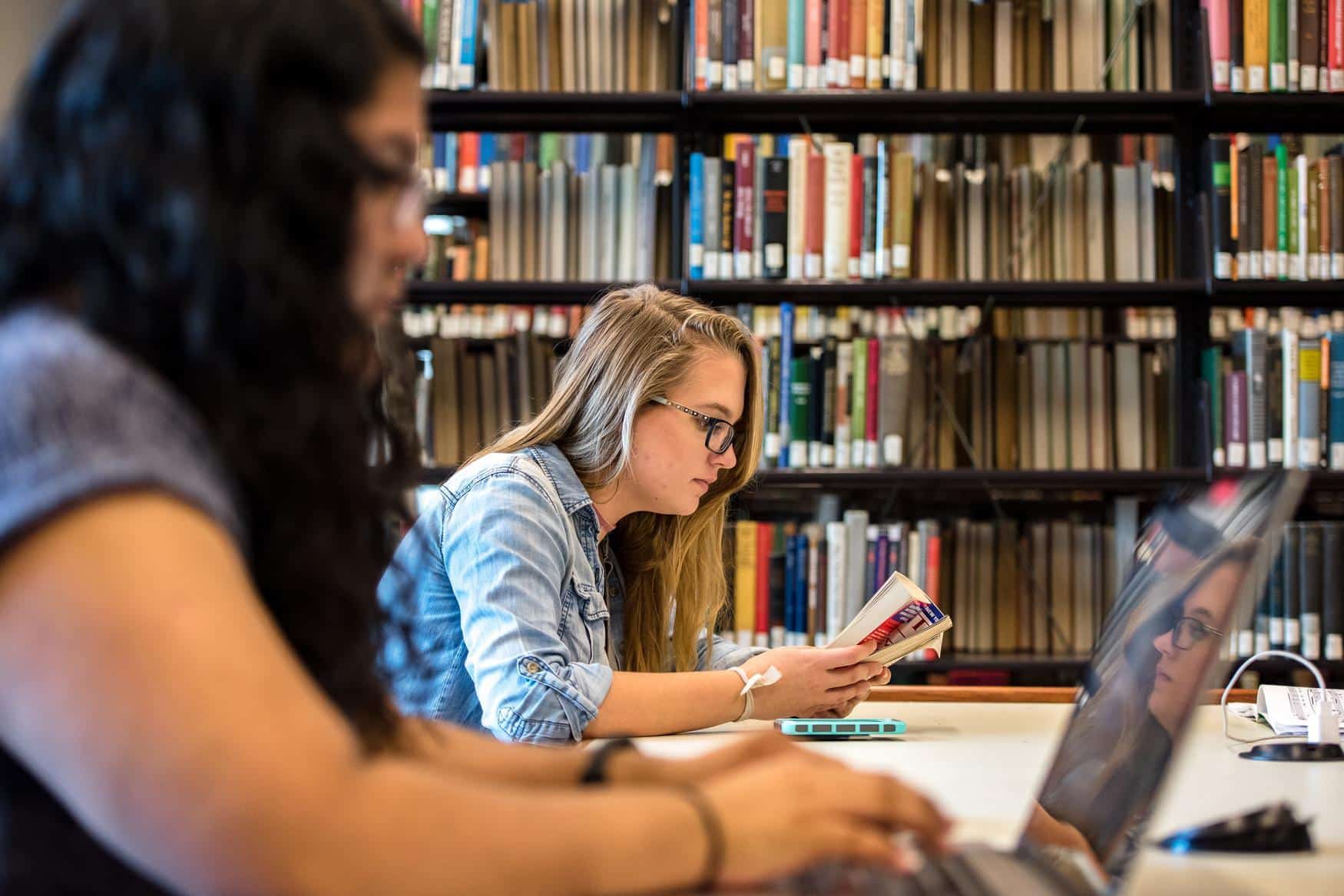 Two women sitting at a desk in a library. One woman is reading a book in the background, and the foreground is a blurry woman on a laptop.