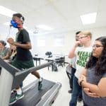 Young man walking on a treadmill with a face mask. A group of students are next to him observing his progress.