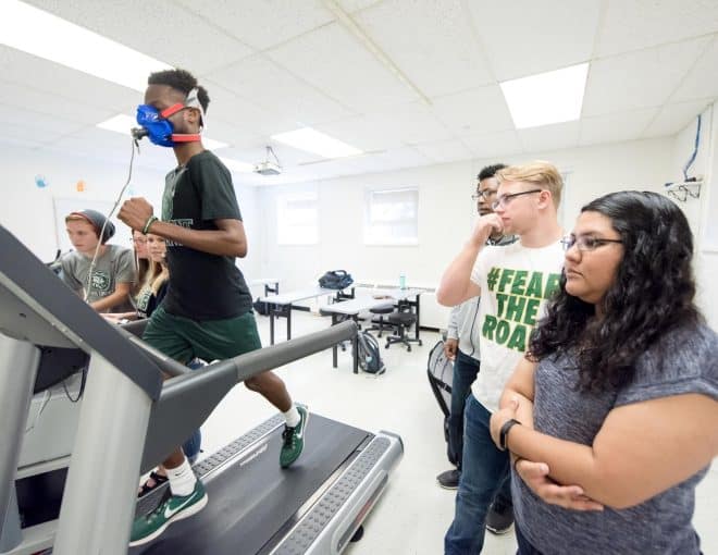 Young man walking on a treadmill with a face mask. A group of students are next to him observing his progress.