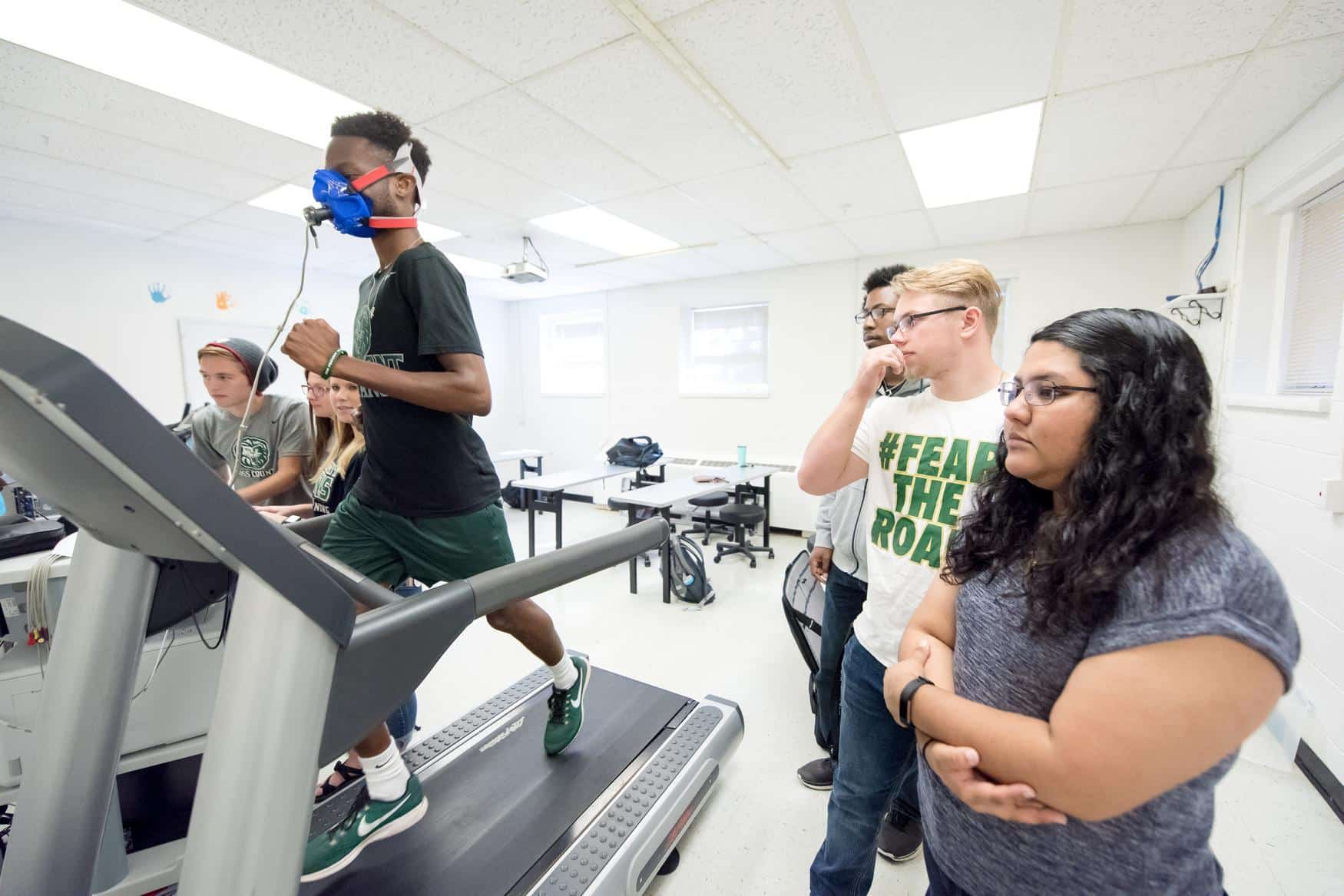 Young man walking on a treadmill with a face mask. A group of students are next to him observing his progress.