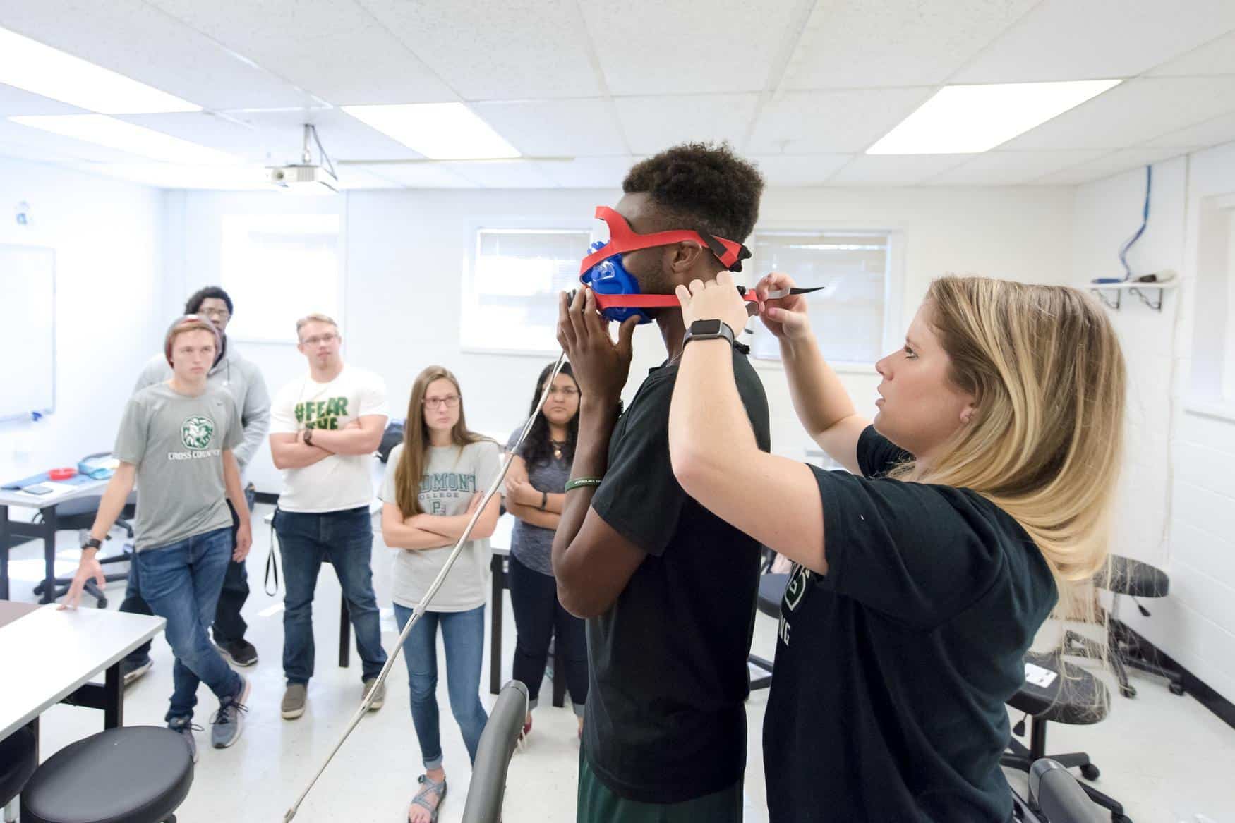 Two students demonstrate medical face mask equipment, while a group of other students watch.