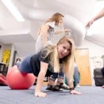 Smiling and laughing woman in the push up position with her feet balancing on a red yoga ball. Other students are laughing and smiling around her.