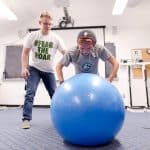 Student doing arm and balance exercises on a large blue yoga ball. Another student stabilizes them and acts as a spotter.
