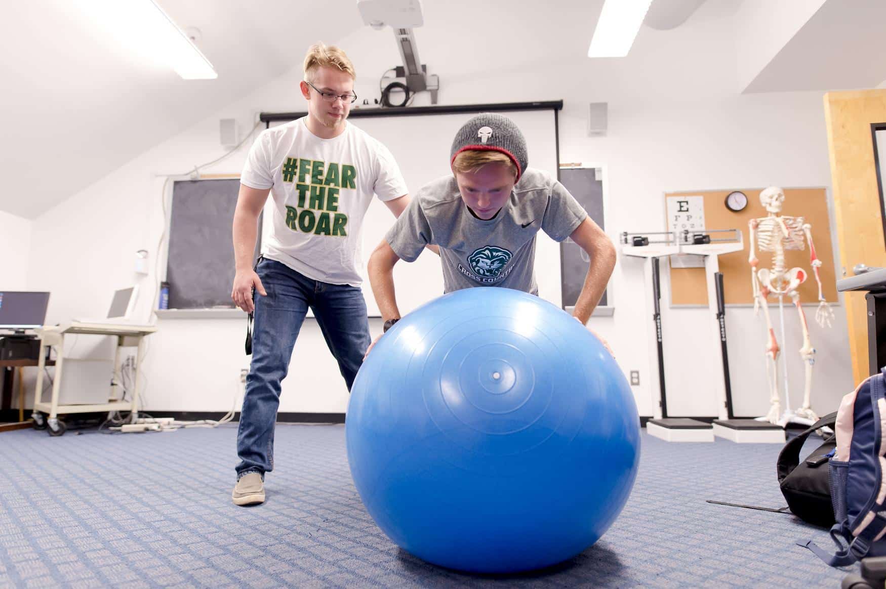 Student doing arm and balance exercises on a large blue yoga ball. Another student stabilizes them and acts as a spotter.