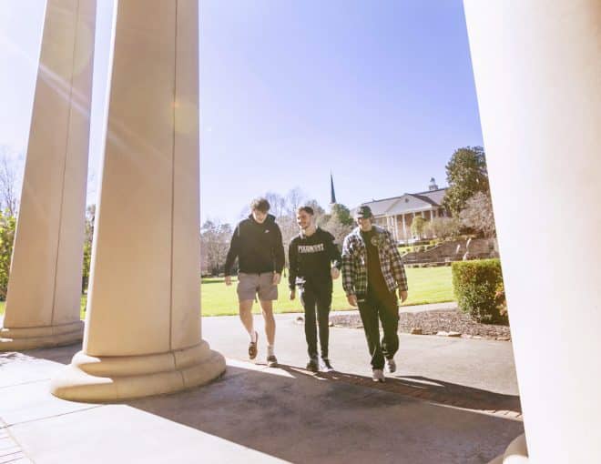 Three people walk together between large columns on a sunny day, with green grass, trees, and a building with a spire in the background.