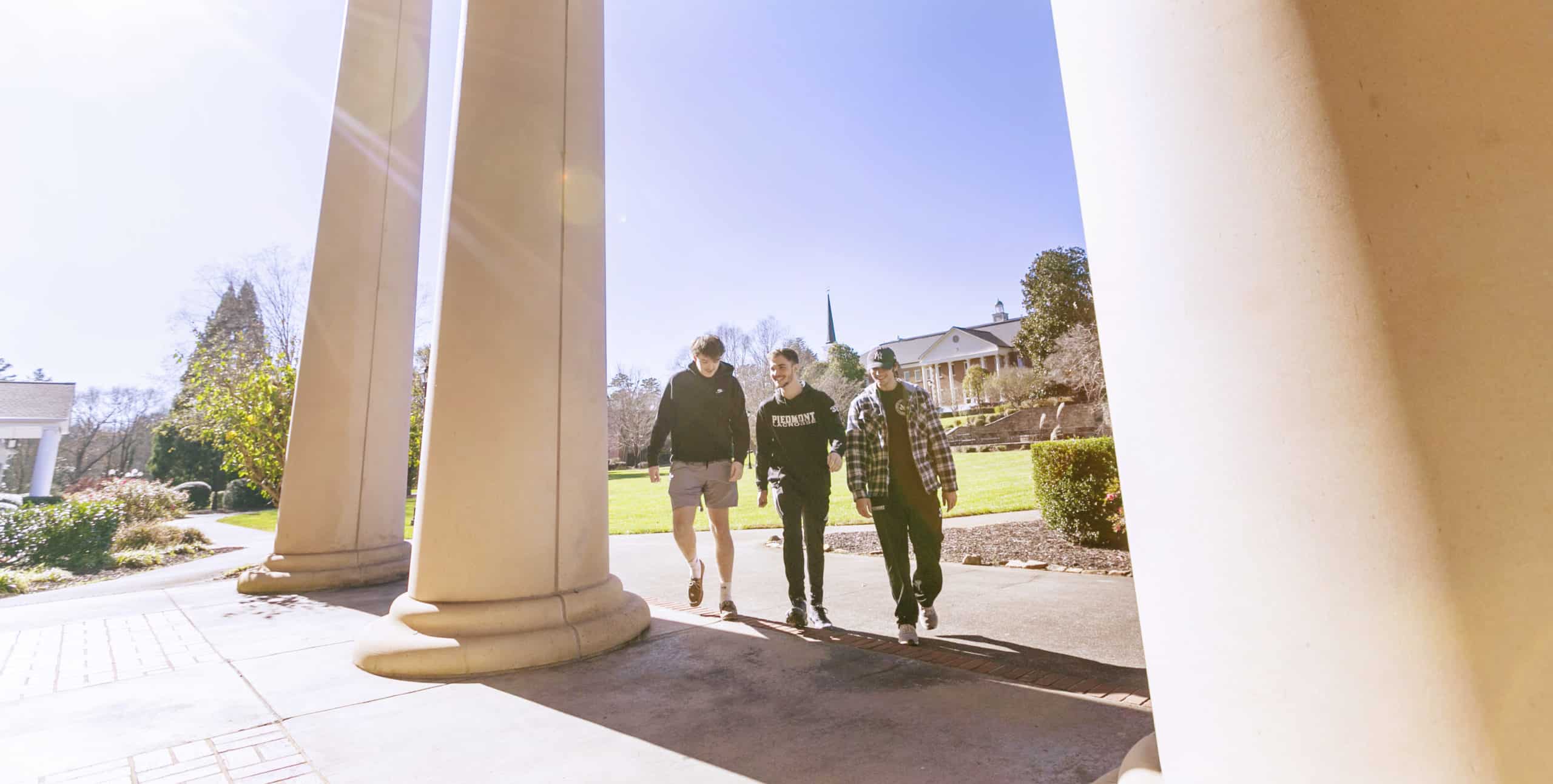 Three people walk together between large columns on a sunny day, with green grass, trees, and a building with a spire in the background.