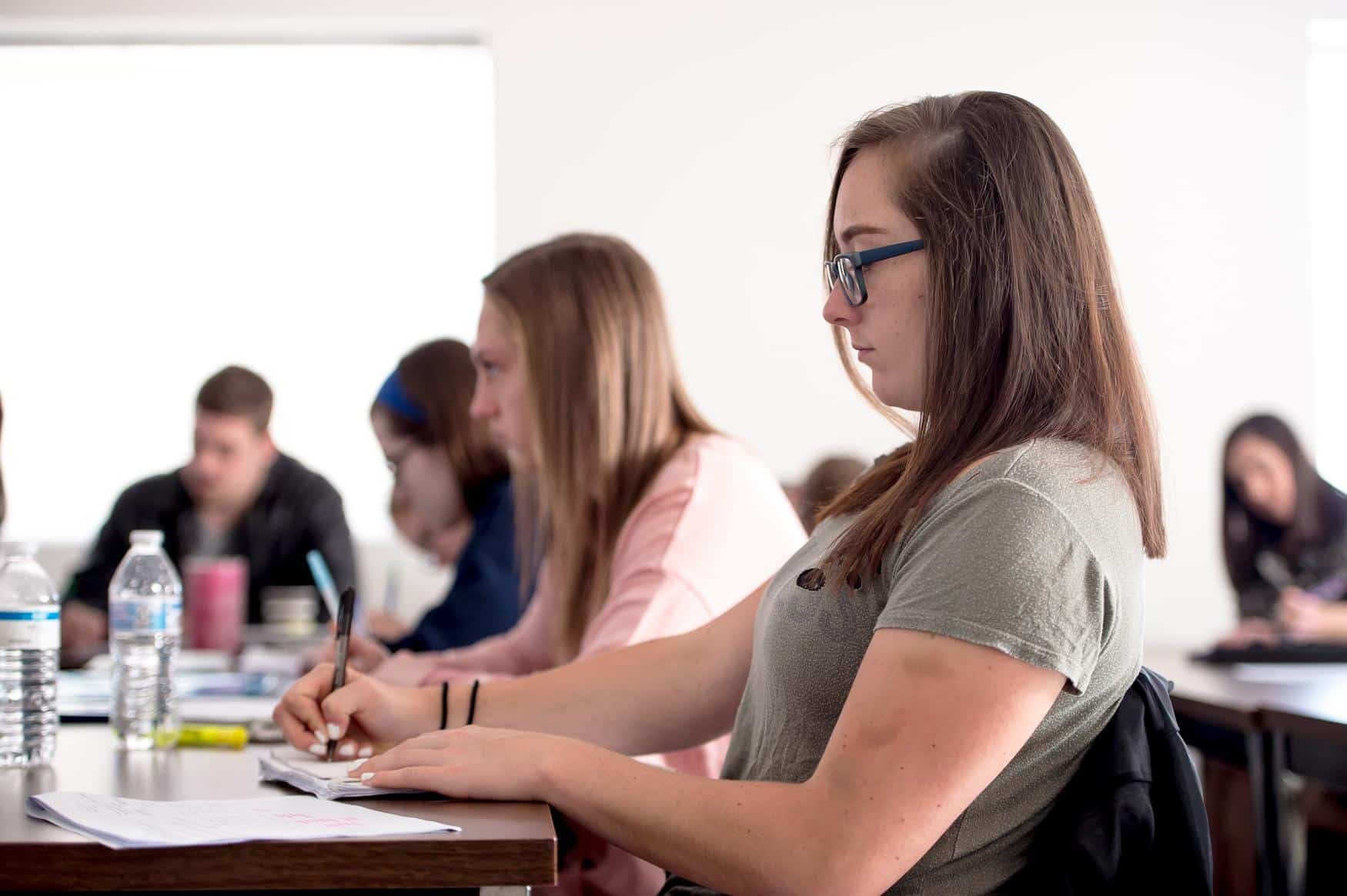 Student with glasses writing notes on a desk. Other students sit beside her but are blurred in the background.