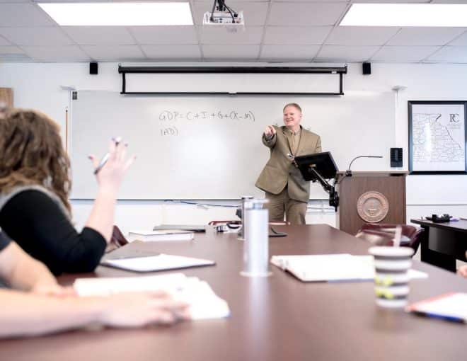 A professor stands at the front of a class, smiling and pointing towards a student with their hand raised in question