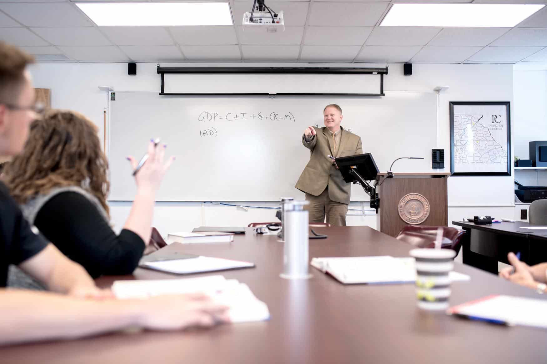 A professor stands at the front of a class, smiling and pointing towards a student with their hand raised in question