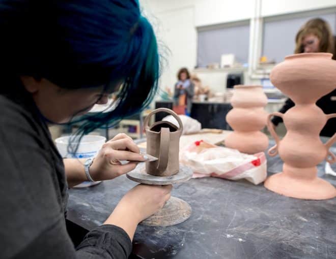 A person with blue hair sculpts a clay object on a stand, while others work on larger pottery pieces in the background in a ceramics studio.