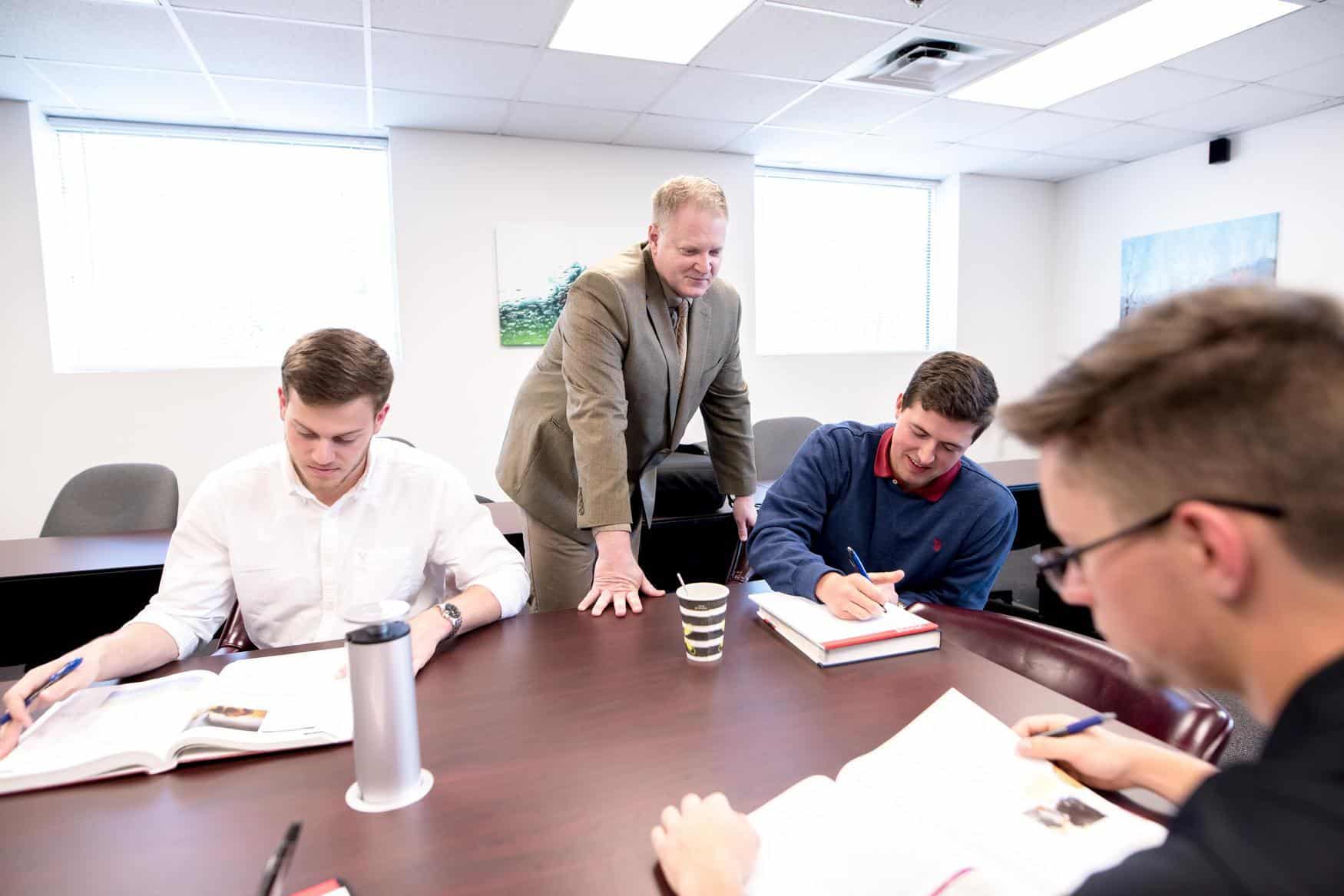 A teacher in a suit stands at a table, engaging with three students who are seated, reading textbooks and taking notes in a bright classroom.