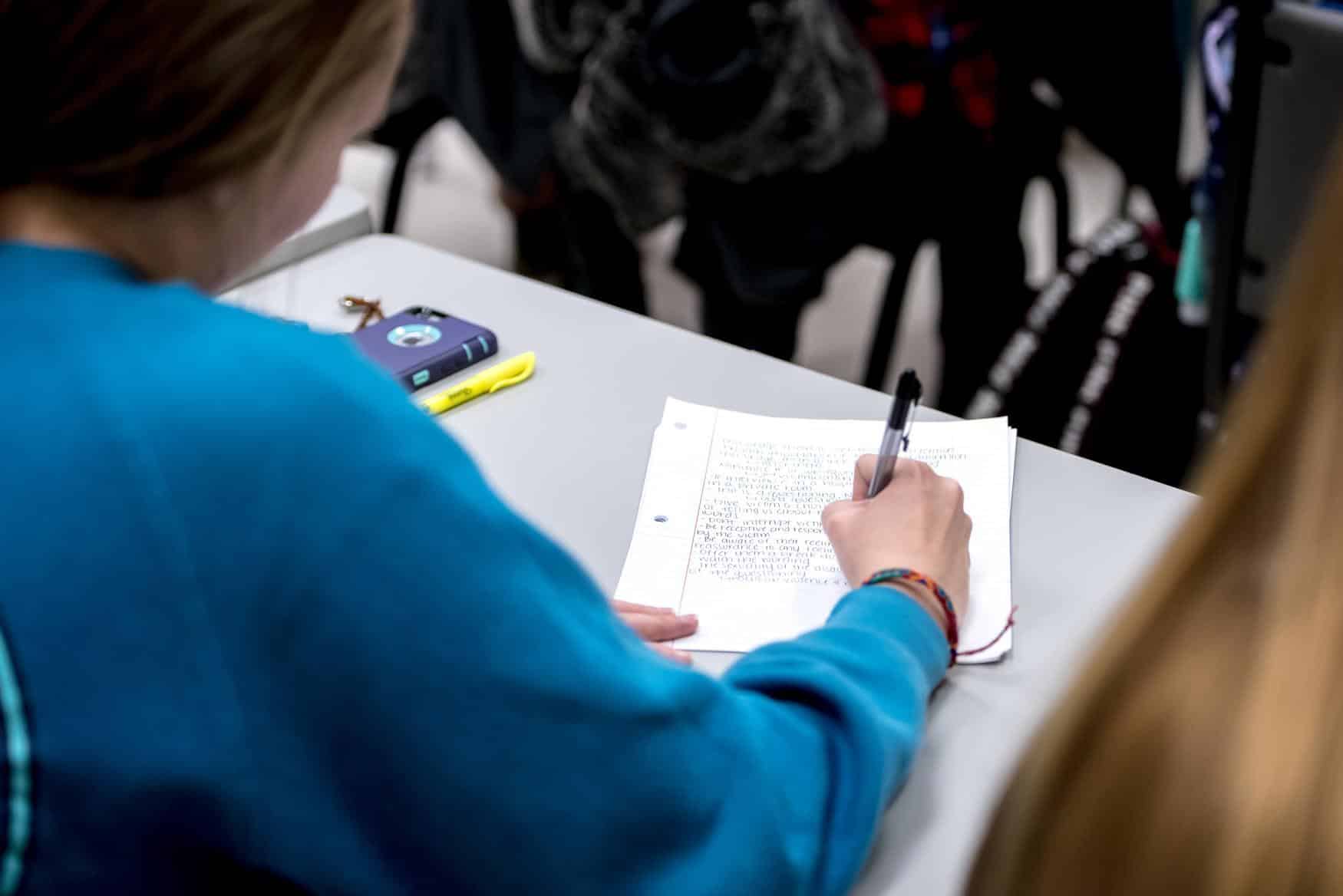 A student wearing a blue sweatshirt writes notes in a notebook with a pen while sitting at a desk. A cell phone, highlighter, and other students are partially visible in the background.