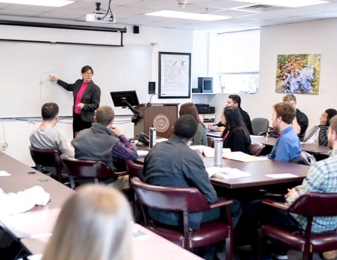 A teacher stands at the front of a classroom, pointing at a whiteboard while students sit at tables, facing her and listening attentively. The classroom has bright windows and various wall decorations.