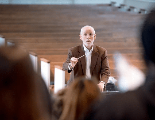 An older man with glasses and a beard, wearing a brown blazer, stands conducting a group with a baton in hand, facing an audience in a modern, wood-paneled room.